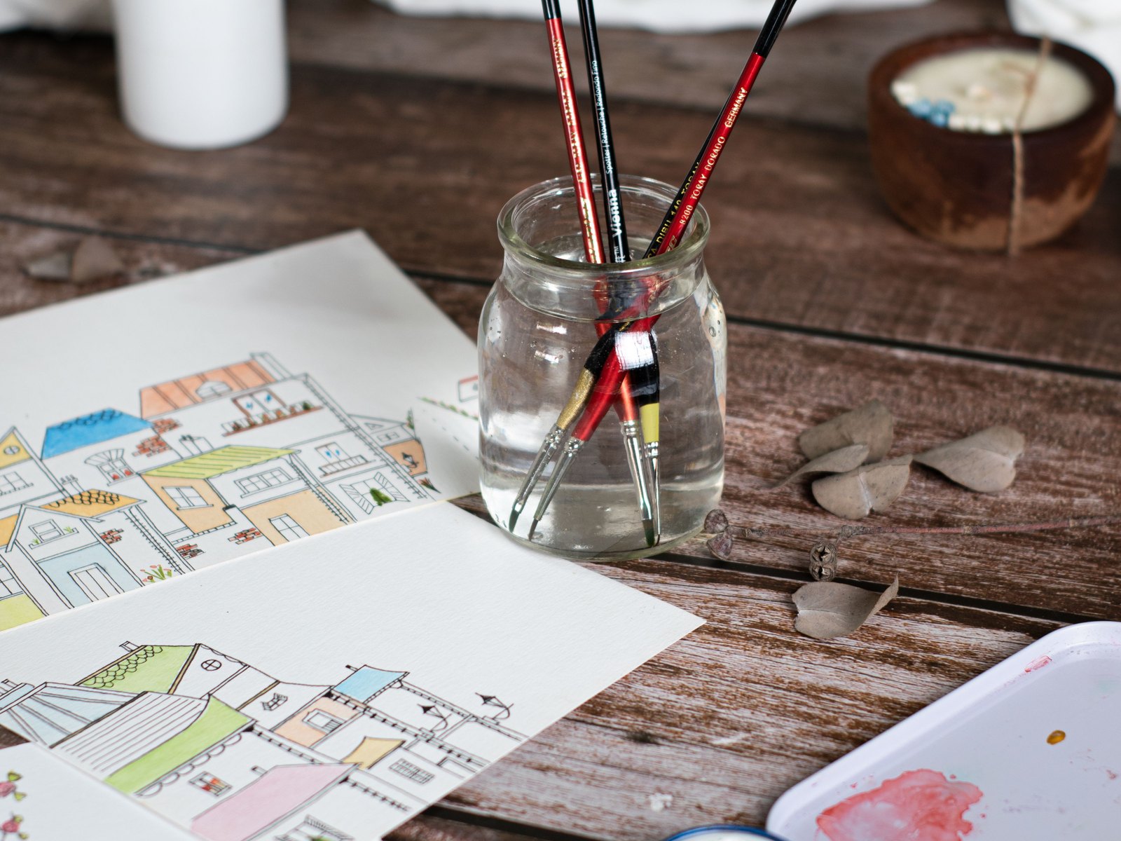 木の机の上の水彩の道具。ガラスの瓶に筆が立ててあり、まちをかいた絵と絵の具のパレットが並んでいる