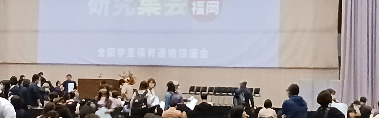 全国学童保育研究集会の会場。大きなスクリーンの前に、大勢の参加者が座っている