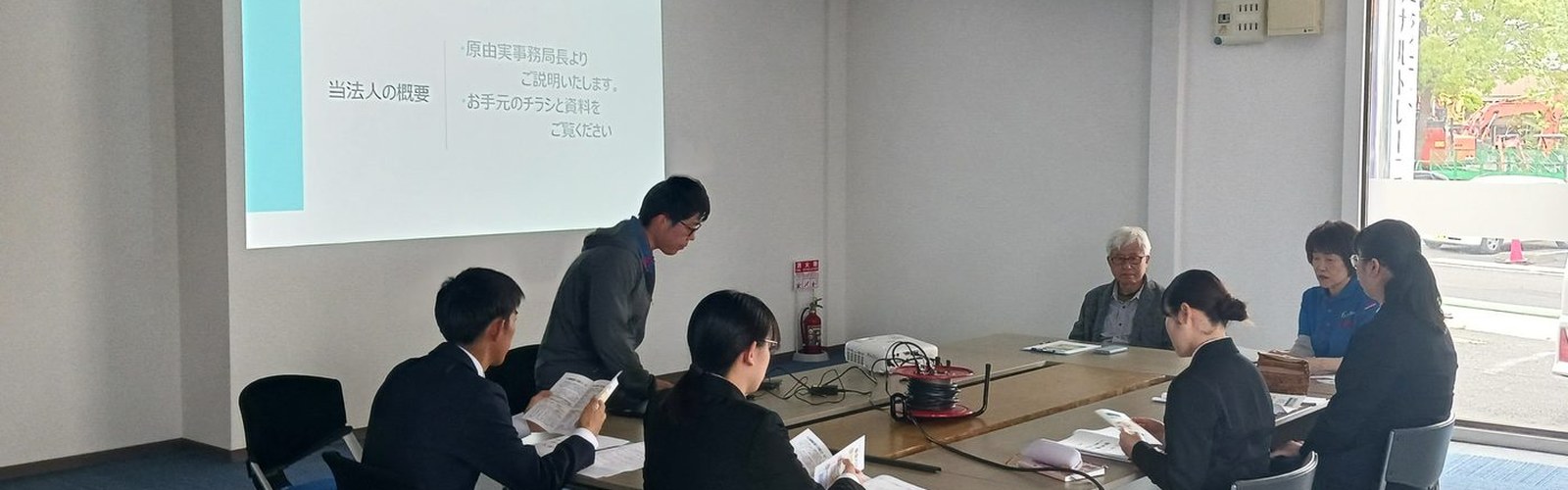 法人の説明会。スクリーンに資料を映し、机を囲んで話を聞いている