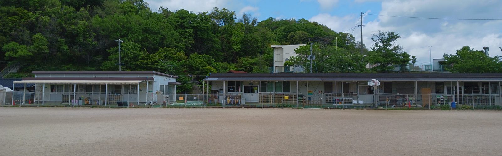 校庭ごしに見える、平屋の児童クラブの建物。奥は緑の山と青空