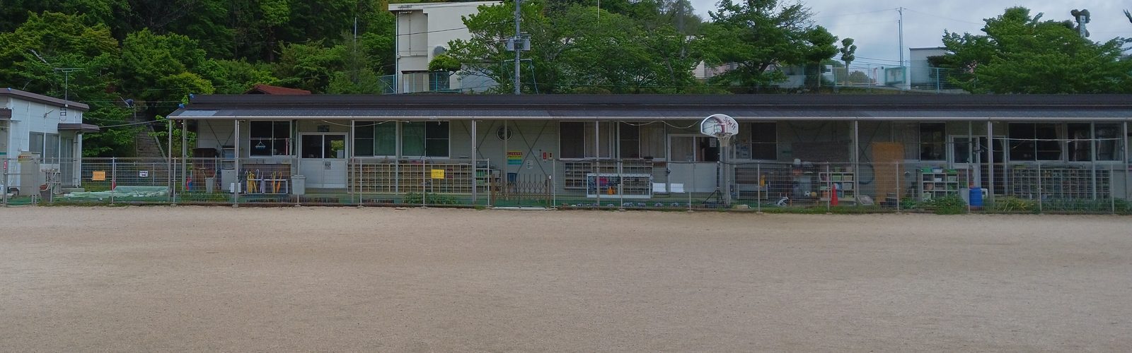 ながおキッZ児童クラブの外観。細長い平屋のプレハブが横に長く続き、奥に緑の山