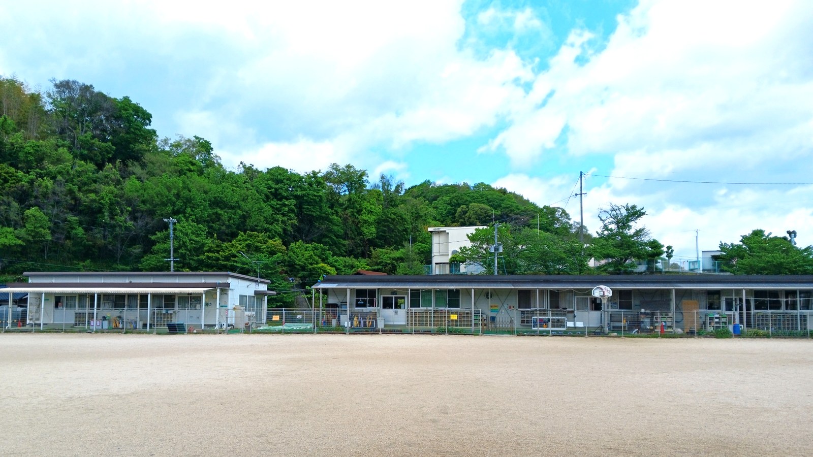 土のグラウンドの向こうに立つ、平屋のプレハブ2棟。右が横に長い棟、左が小さい棟で、うしろに緑の木がしげる山が見える