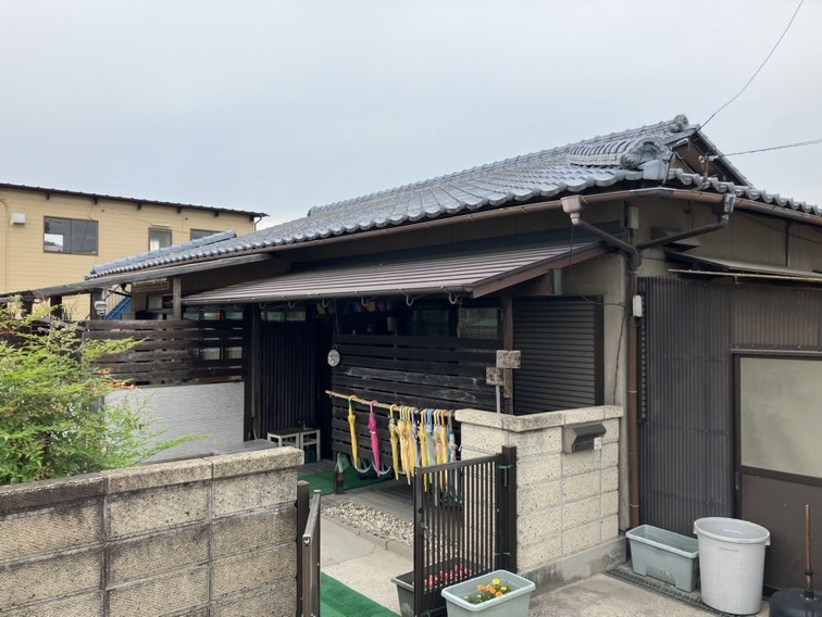 かわら屋根の平屋の民家。玄関の軒下に色とりどりのかさが干してあり、手前にブロック塀と黒い門とびらがある
