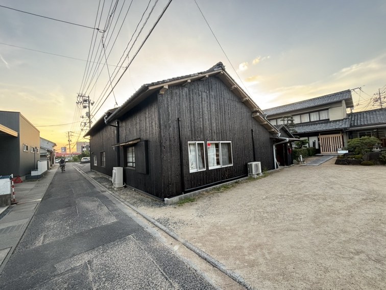 焼き板のような黒い板壁の平屋の建物。三角屋根で道路の角に立ち、となりにかわら屋根の日本家屋がならんでいる