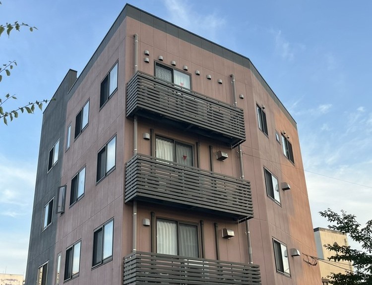 うすいピンク色の外壁の4階建ての建物を、下から見上げたところ。各階に横板のさくのベランダがならんでいる