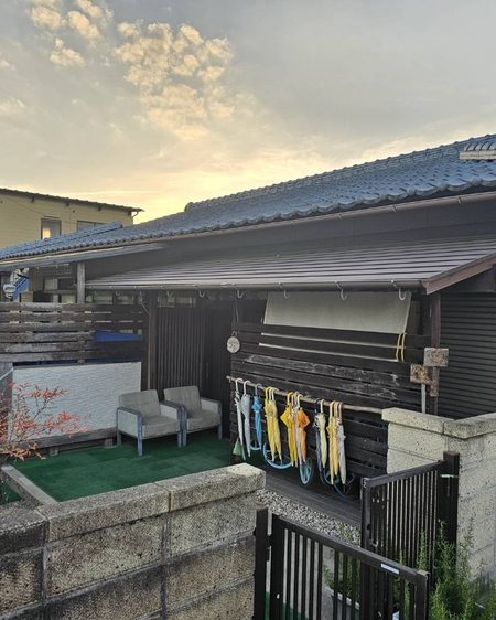 民家の施設の裏庭。人工芝とウッドデッキ、軒下に干された黄色や青のカッパ