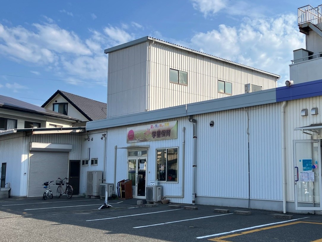 駐車場に面した白い波板の平屋の建物。「中島学童保育」と書かれた横長の看板とガラスの入口があり、その奥に白い2階建ての建物が立っている