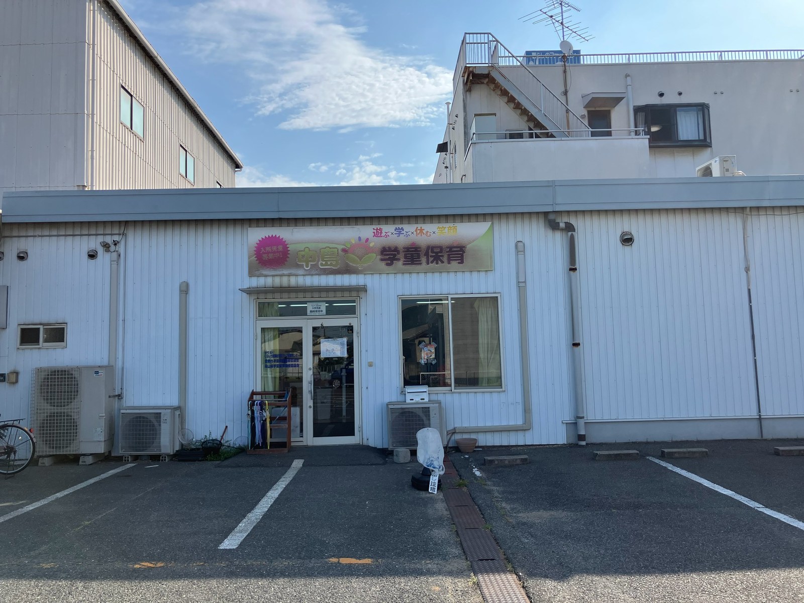 同じ建物の入口を正面から見たところ。「中島学童保育 遊ぶ×学ぶ×休む×笑顔」の看板の下にガラスの引き戸があり、わきに傘立てと何本かのかさが置かれている