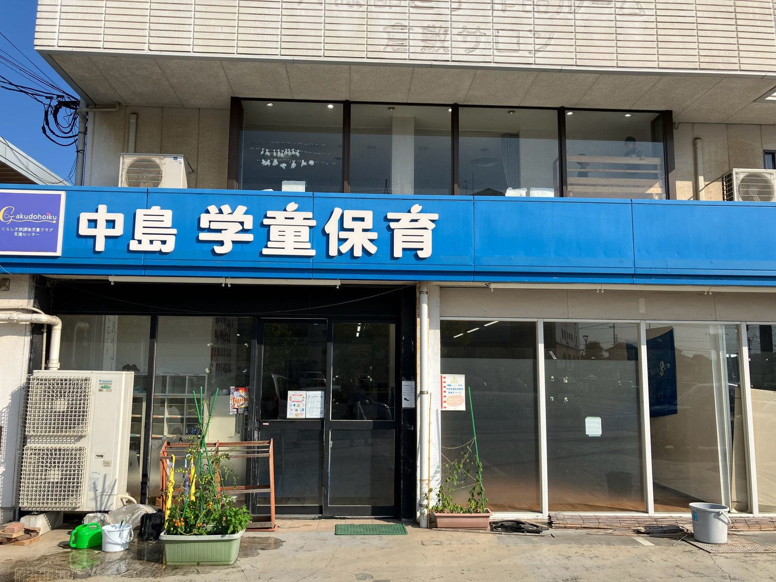 2階建ての建物。1階の上に「中島 学童保育」と白い文字で書かれた青い大きな看板がかかり、その下にガラスの戸と、ミニトマトなどのプランターがならんでいる