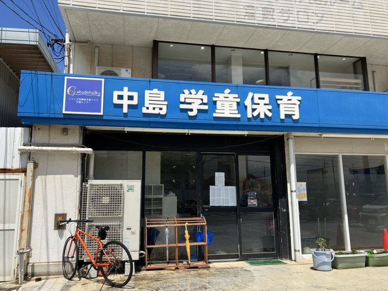 同じ建物の入口に近づいたところ。青い看板の下のガラス戸の前に、オレンジ色の自転車と傘立て、鉢植えの草花が置かれている