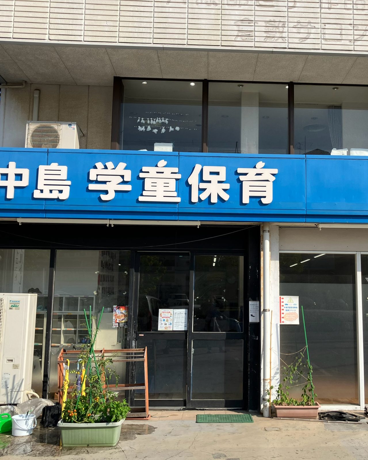 中島学童保育の入口。青い看板の下のガラス戸と、鉢植えの草花