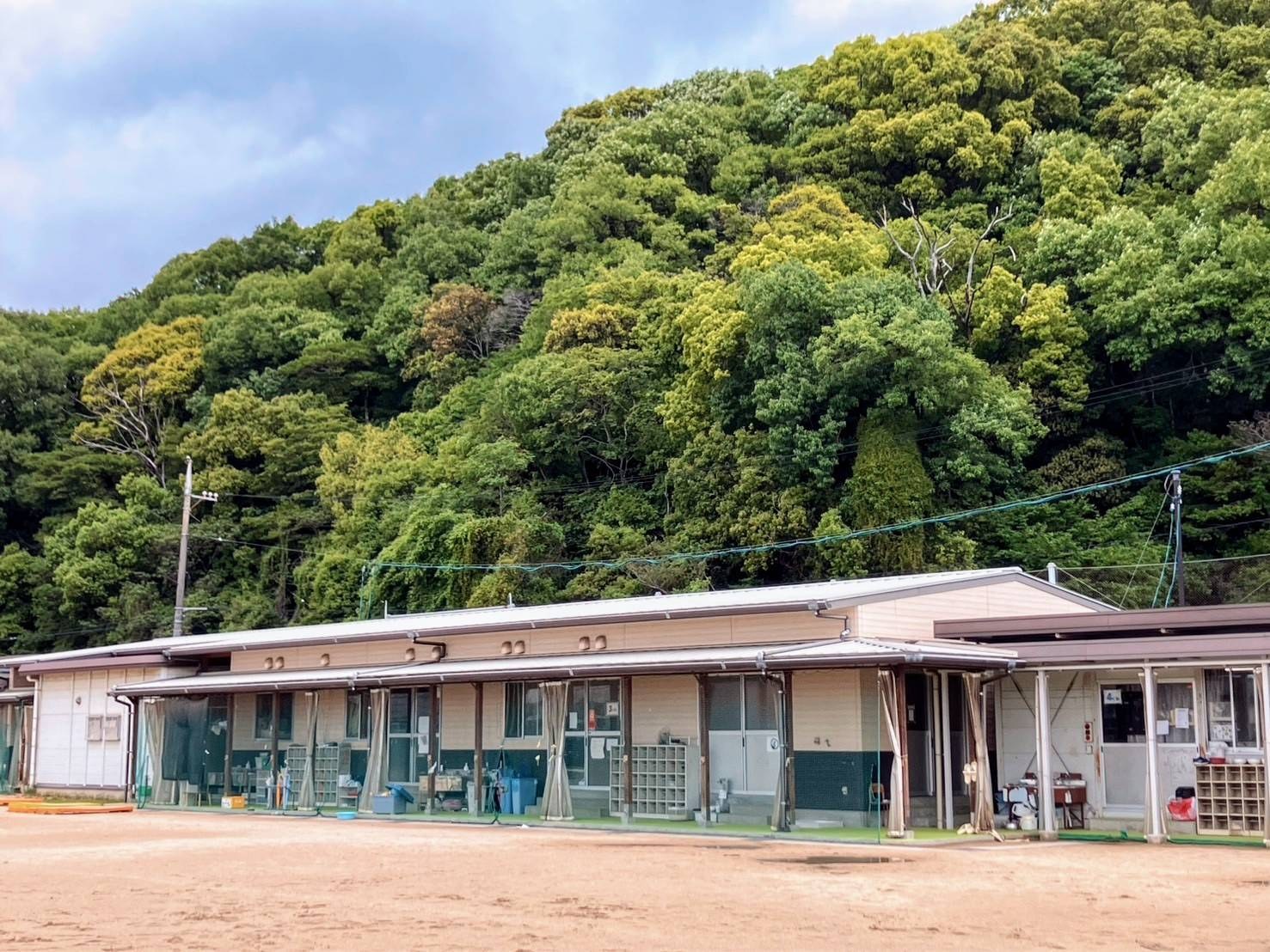 グラウンドの向こうに立つ、横に長い平屋のプレハブ2棟。軒下の通路に下駄箱や道具がならび、うしろに緑の木がしげる山がせまっている