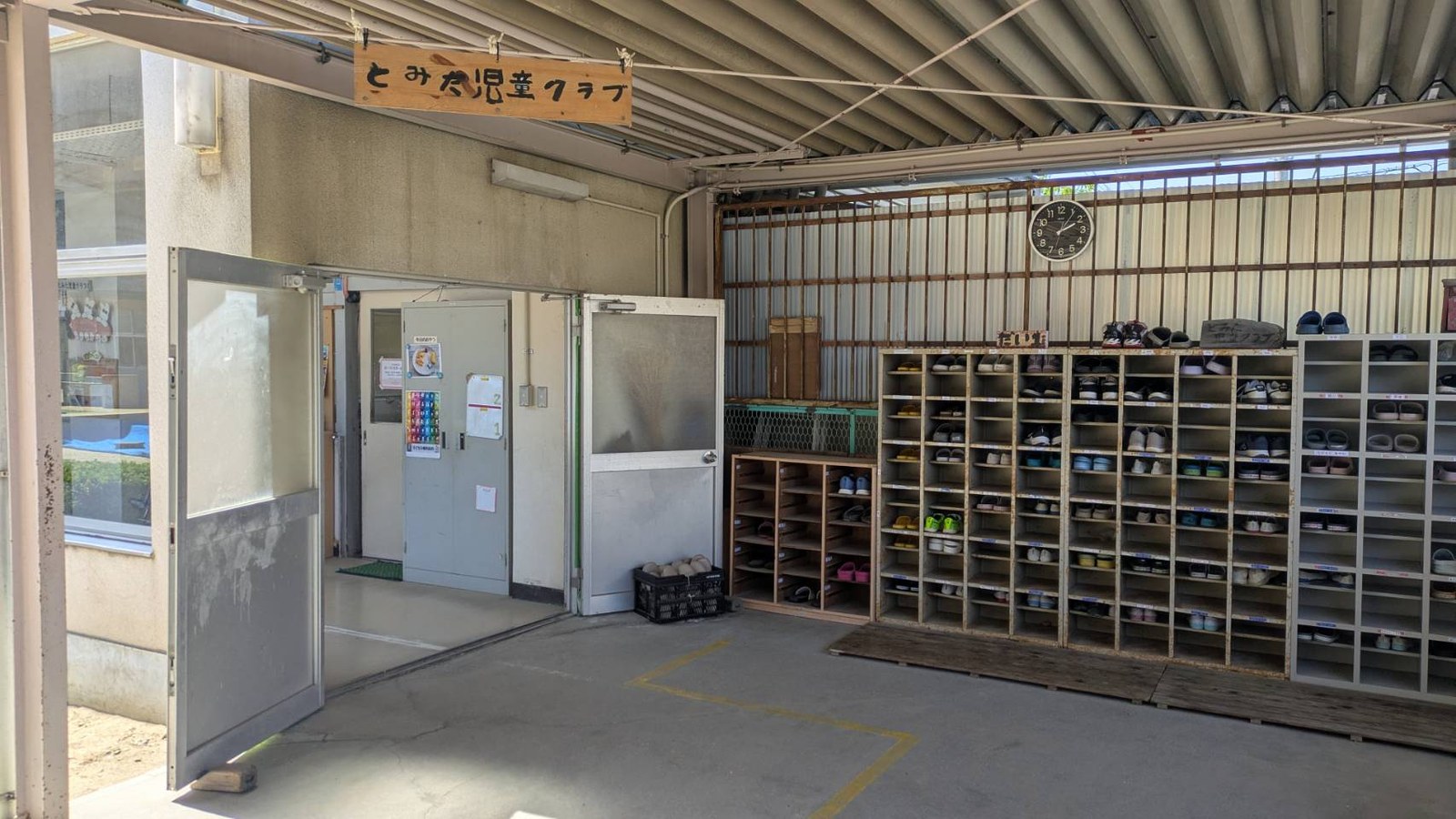 校舎の入口。「とみた児童クラブ」と手書きされた木の看板が下がり、その奥に開いた扉と、たくさんのくつが入った下駄箱がならんでいる