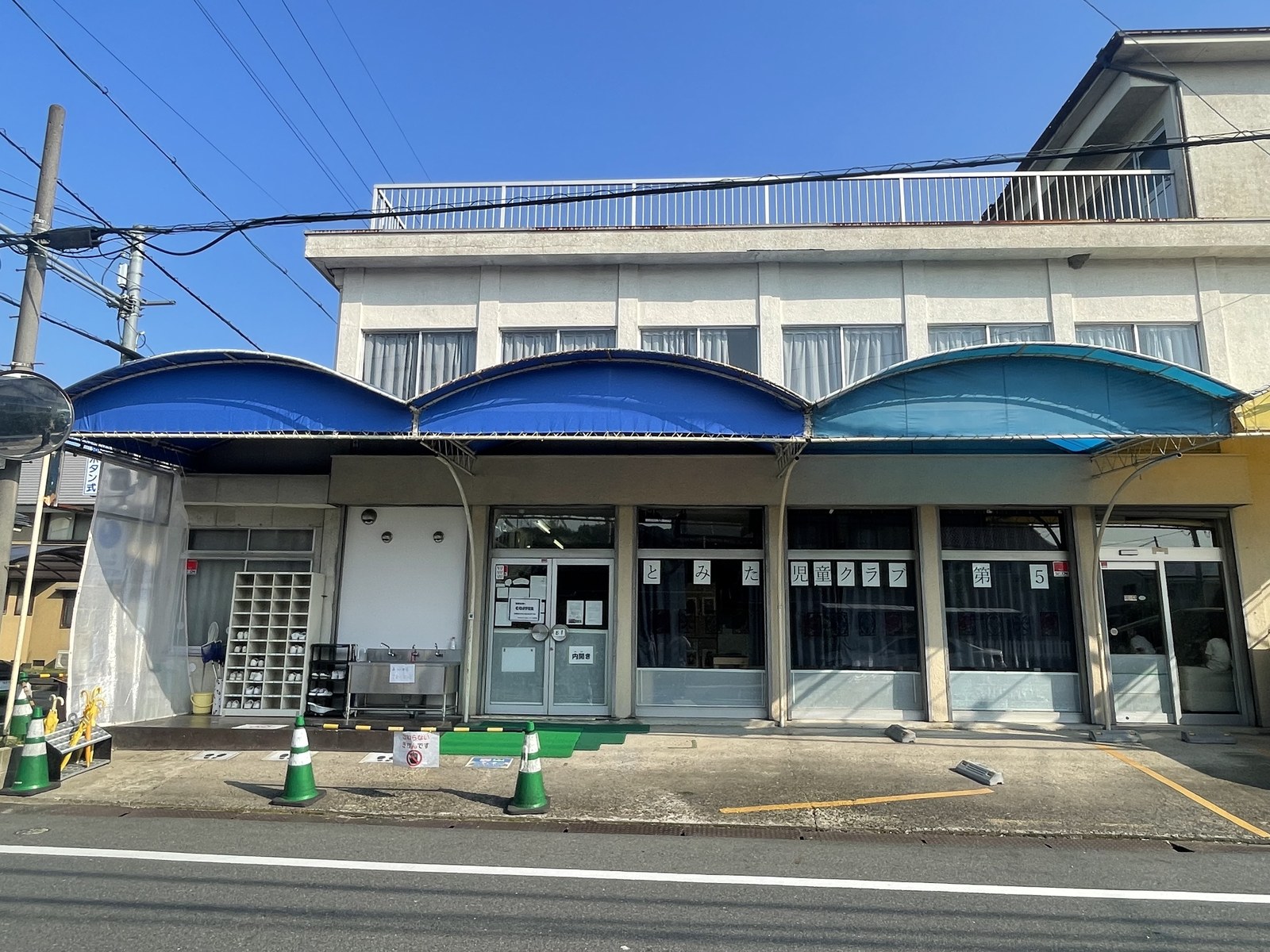 通りに面した2階建ての建物。1階の前に青いアーチ形のひさしが3つならび、ガラス戸のよこの窓に「とみた児童クラブ 第5」と書かれている